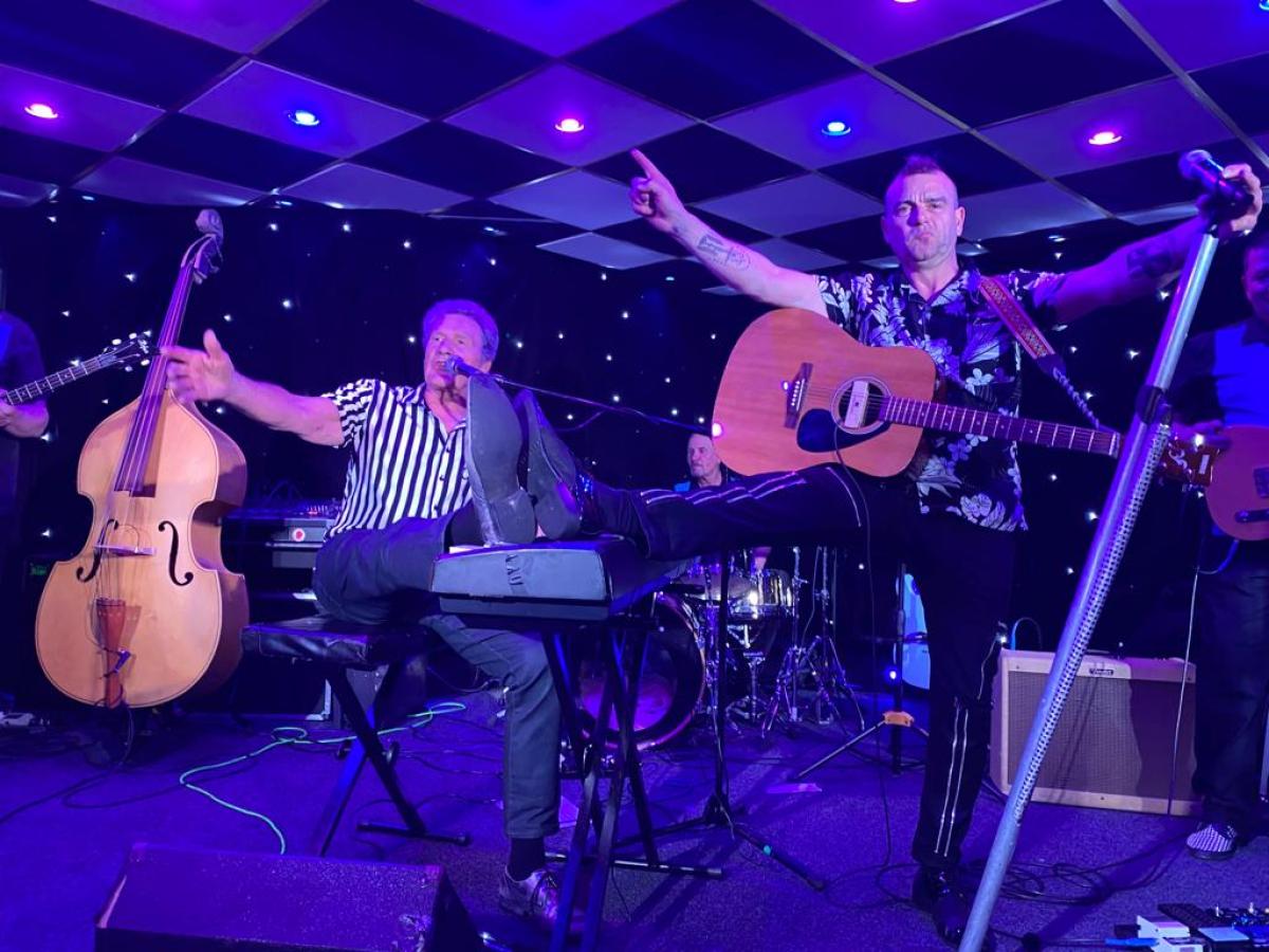 Band performing on stage with colorful lights; man leans on keyboard, another plays guitar.