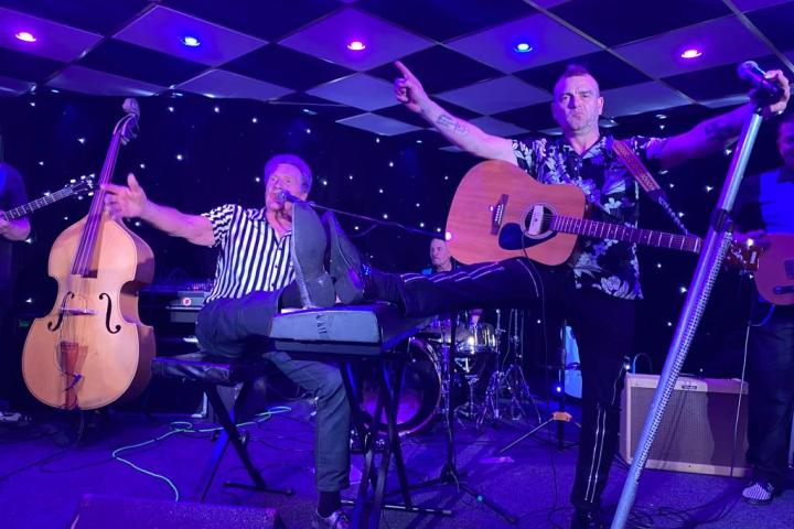 Band performing on stage with colorful lights; man leans on keyboard, another plays guitar.