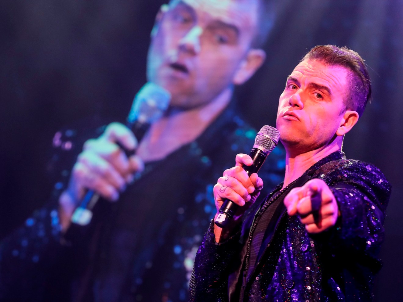 Singer in sparkly jacket holding microphone, pointing, with blurry stage backdrop.