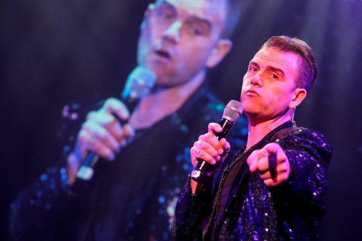 Singer in sparkly jacket holding microphone, pointing, with blurry stage backdrop.