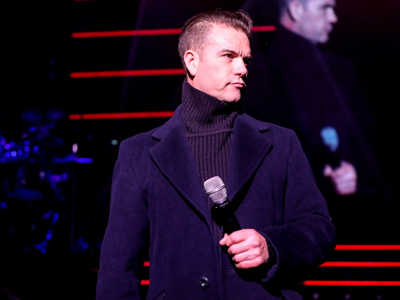 Person in a dark coat holding a microphone on stage with red lights in the background.