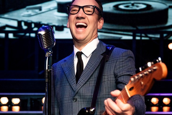 Smiling musician with glasses singing, playing guitar by vintage microphone on stage.