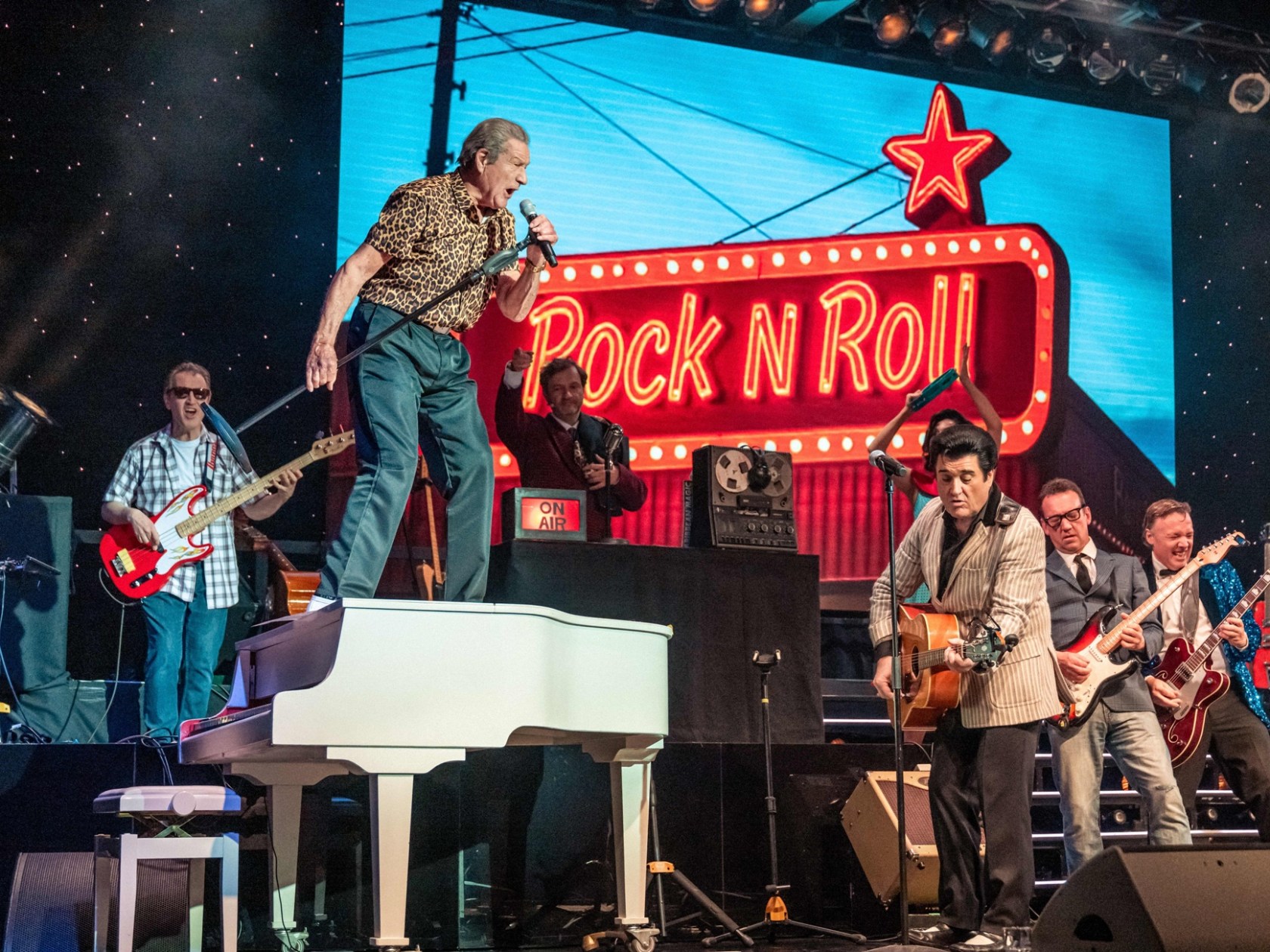 Band performing on stage with rock 'n' roll sign, singer standing on piano.