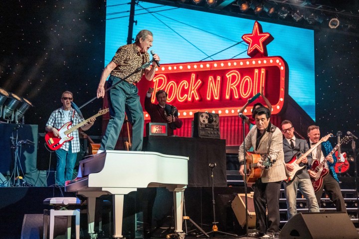 Band performing on stage with rock 'n' roll sign, singer standing on piano.