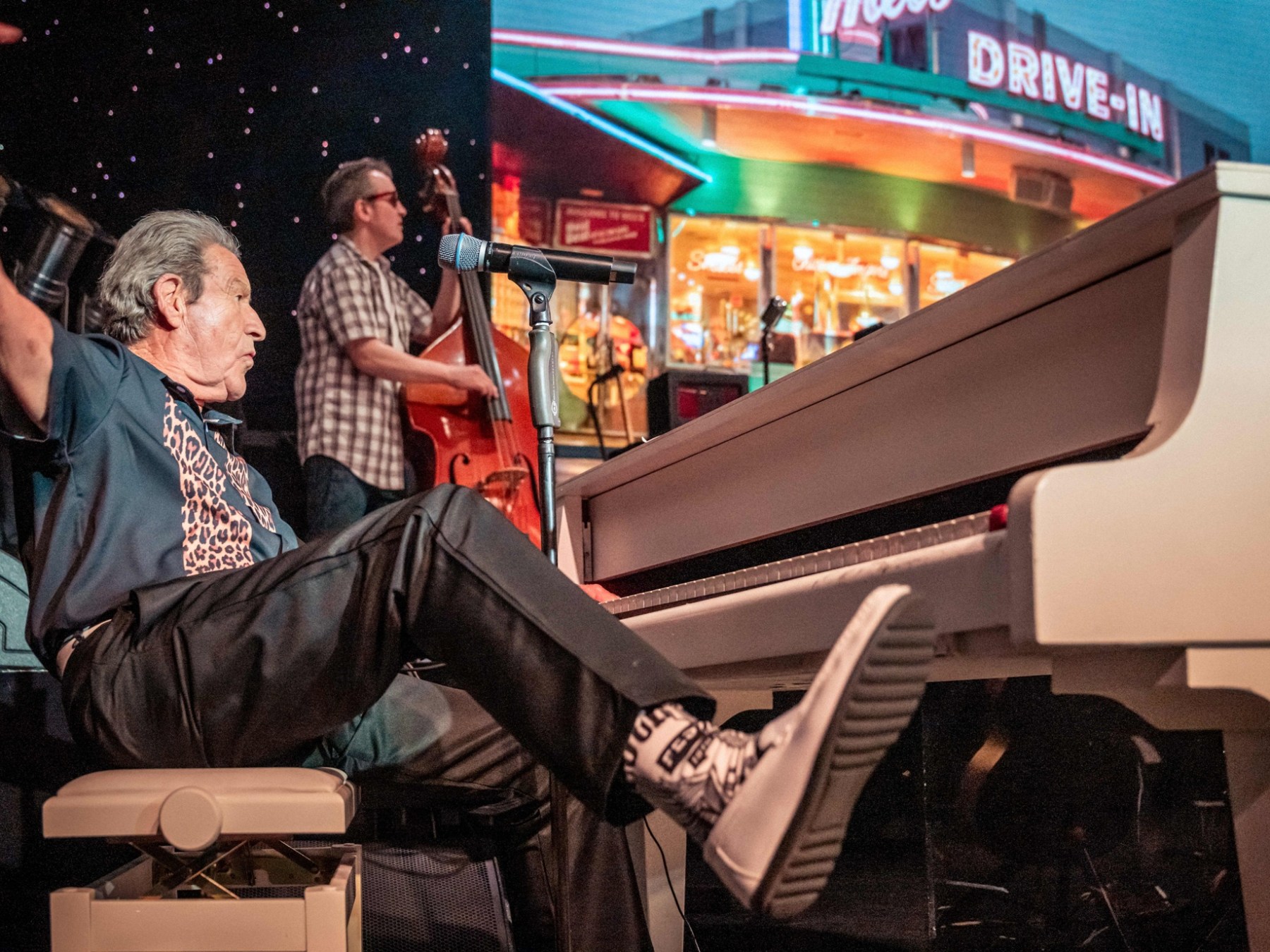 Man playing white piano energetically, foot on keys, with bassist in background by diner sign.