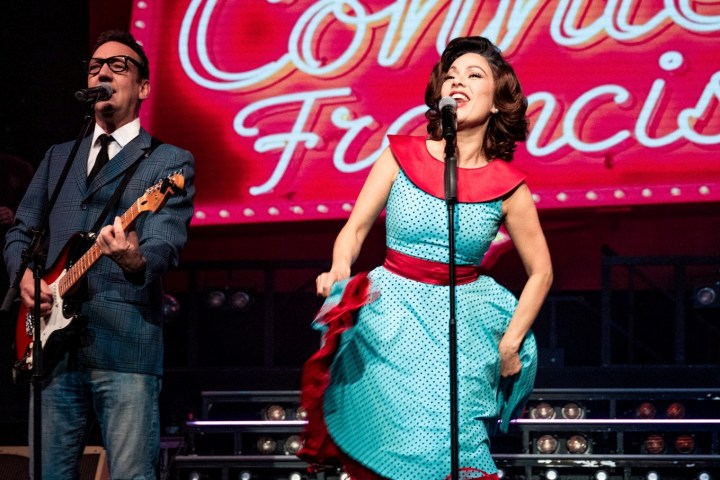 A man playing guitar and a woman singing on stage with 'Connie Francis' sign behind.