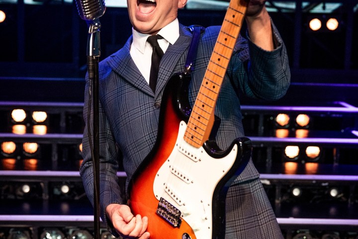 Person in suit passionately playing electric guitar on stage with vintage microphone.