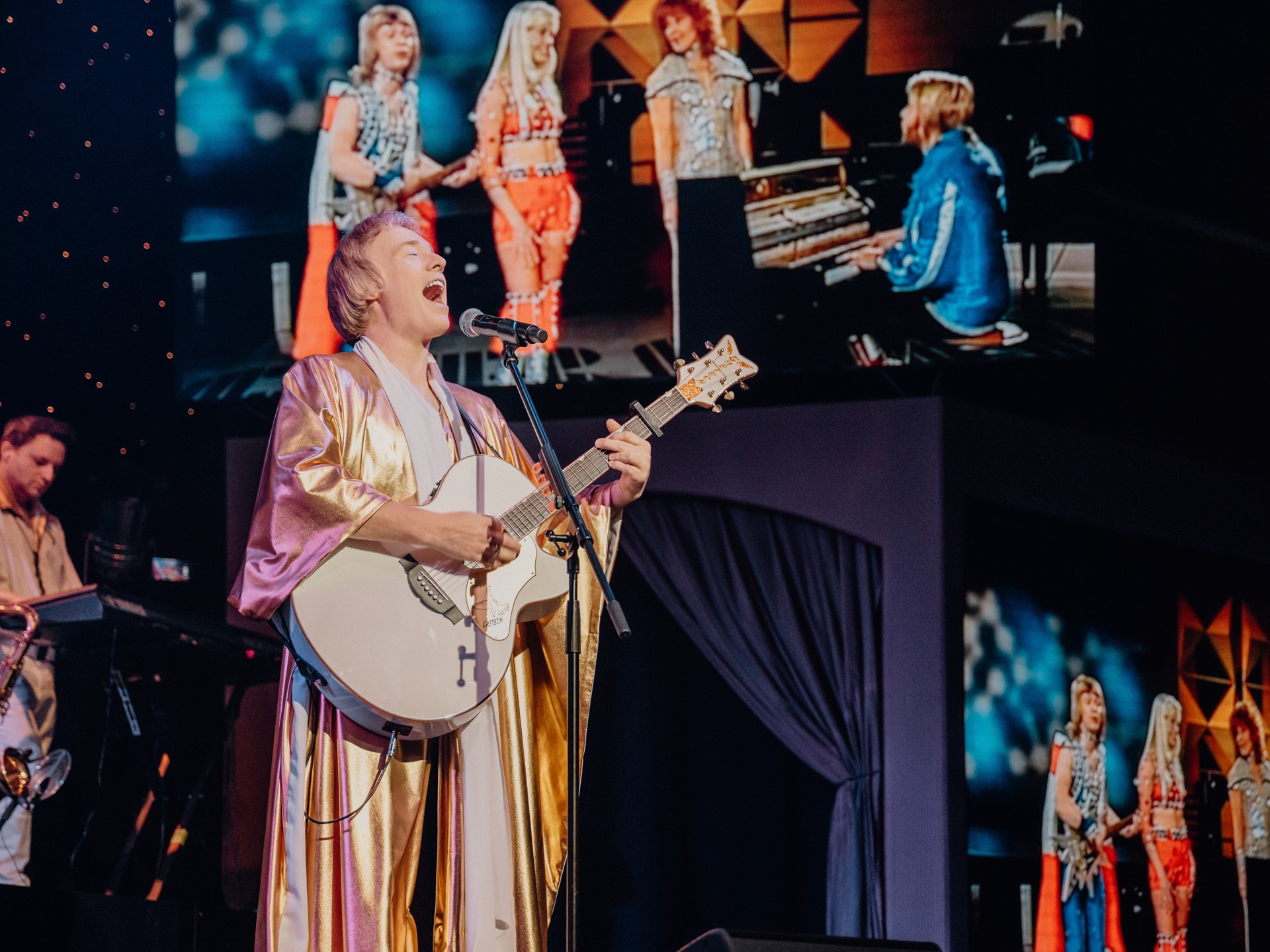 Person in a gold robe playing guitar and singing on stage, with band and video screen behind.