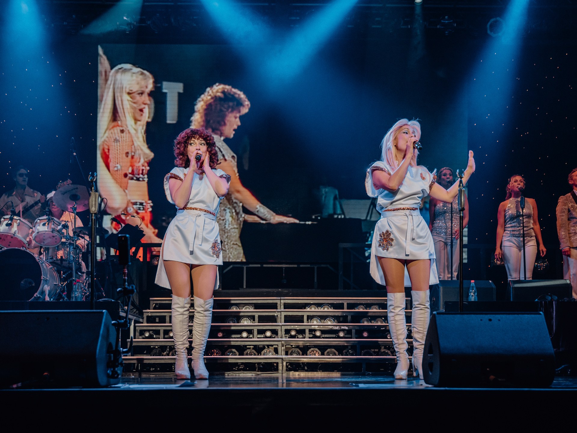 Two female singers performing on stage with a band and a large screen backdrop.