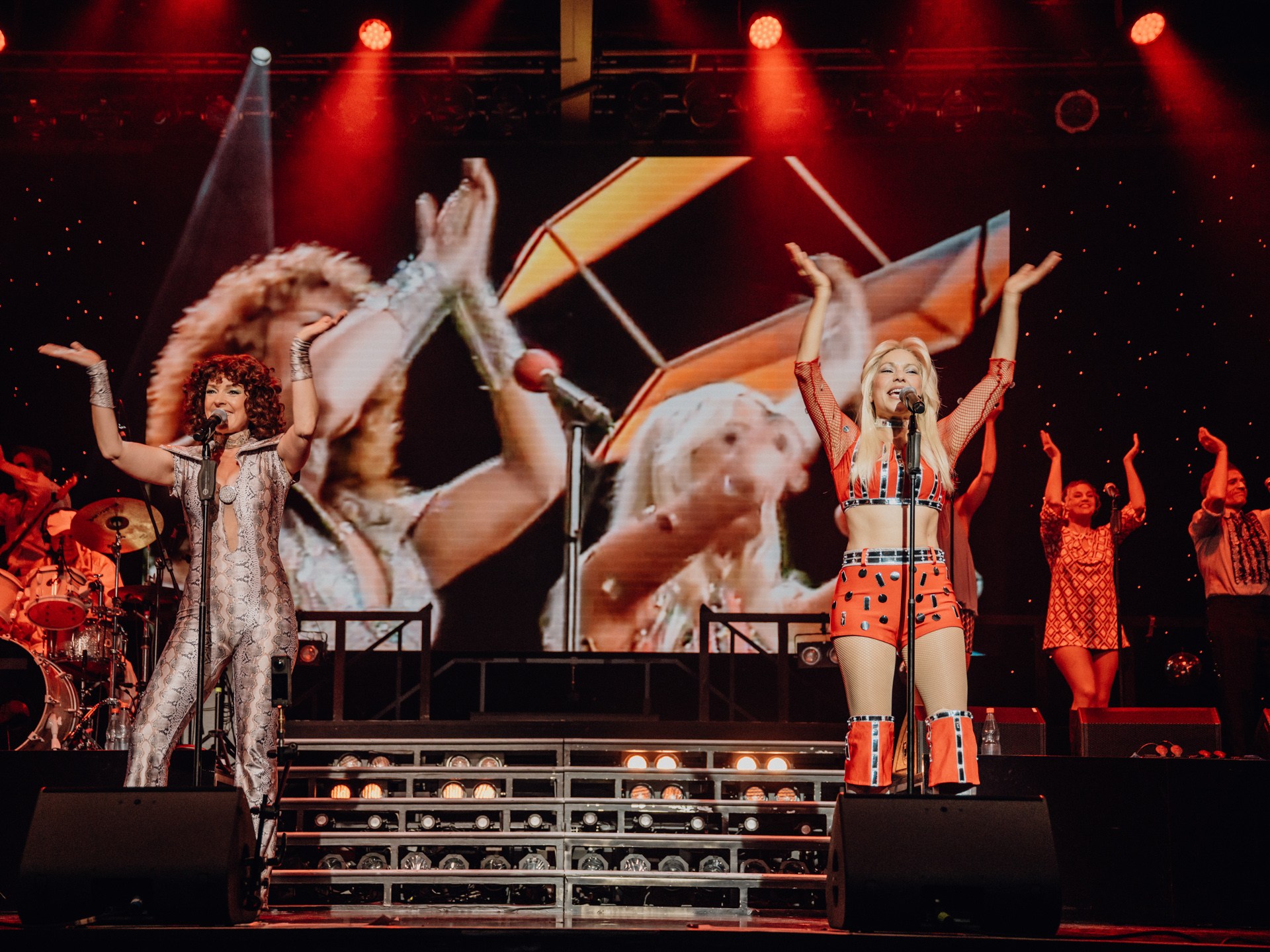 Performers on stage with arms raised, colorful outfits, and large screen displaying performers in background.