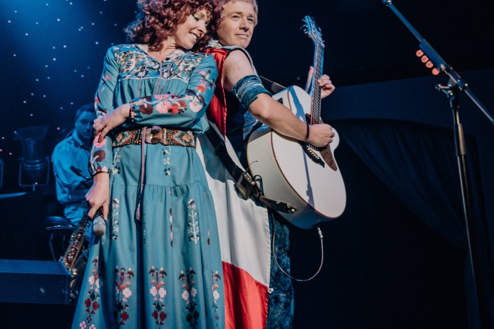 Two musicians on stage, one with a guitar, and the other holding a microphone under blue stage lights.