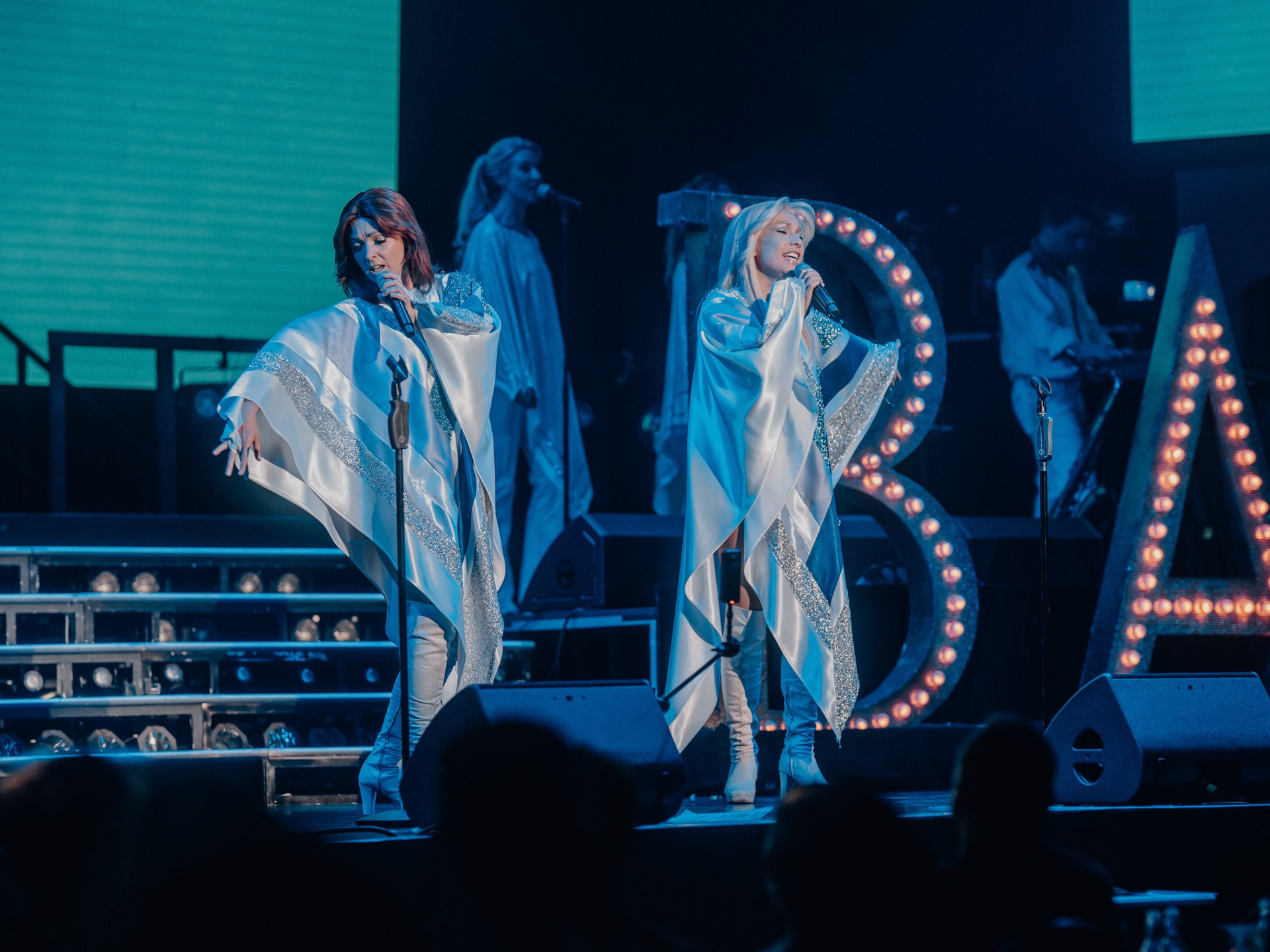 Two women singing on stage with a band and illuminated letters in the background.