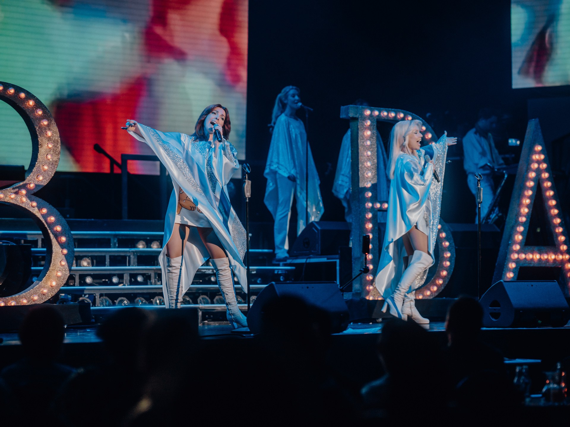 Two performers in matching outfits sing on stage with large lit letters B and A behind them.