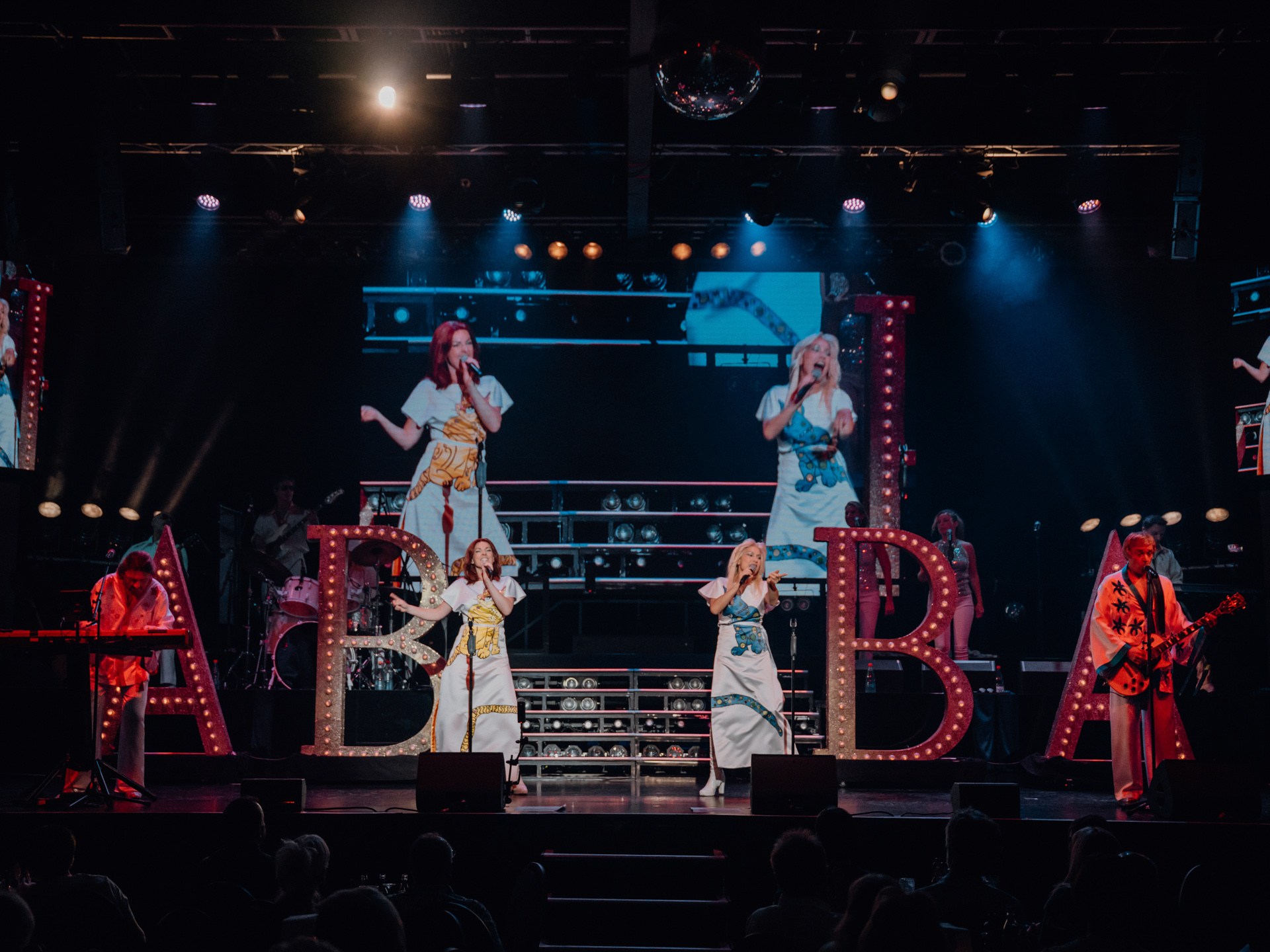 Band in ABBA costumes performing on stage with large lit letters behind them.