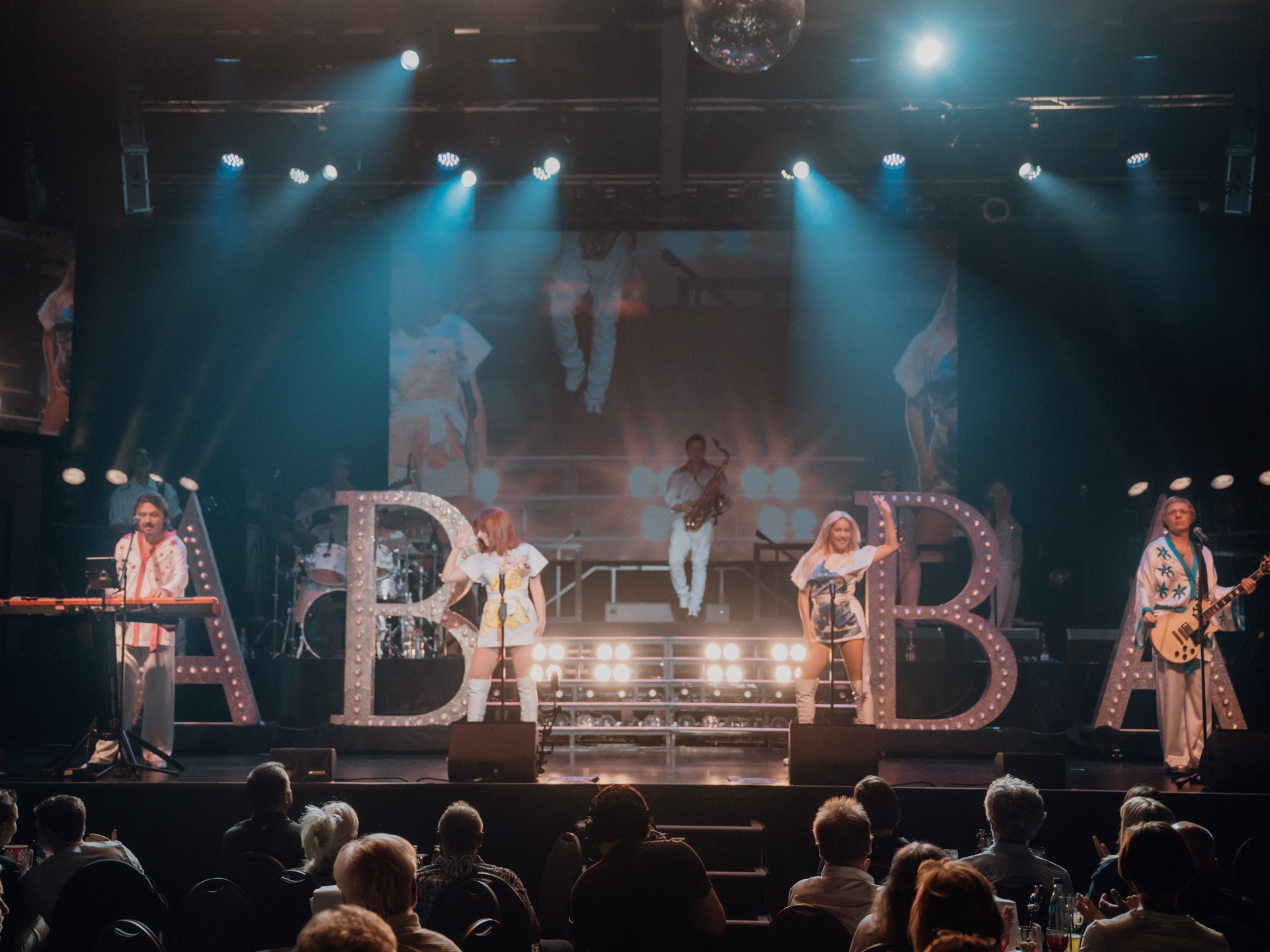 Band performing on stage with large lit letters 'ABBA' and audience watching.