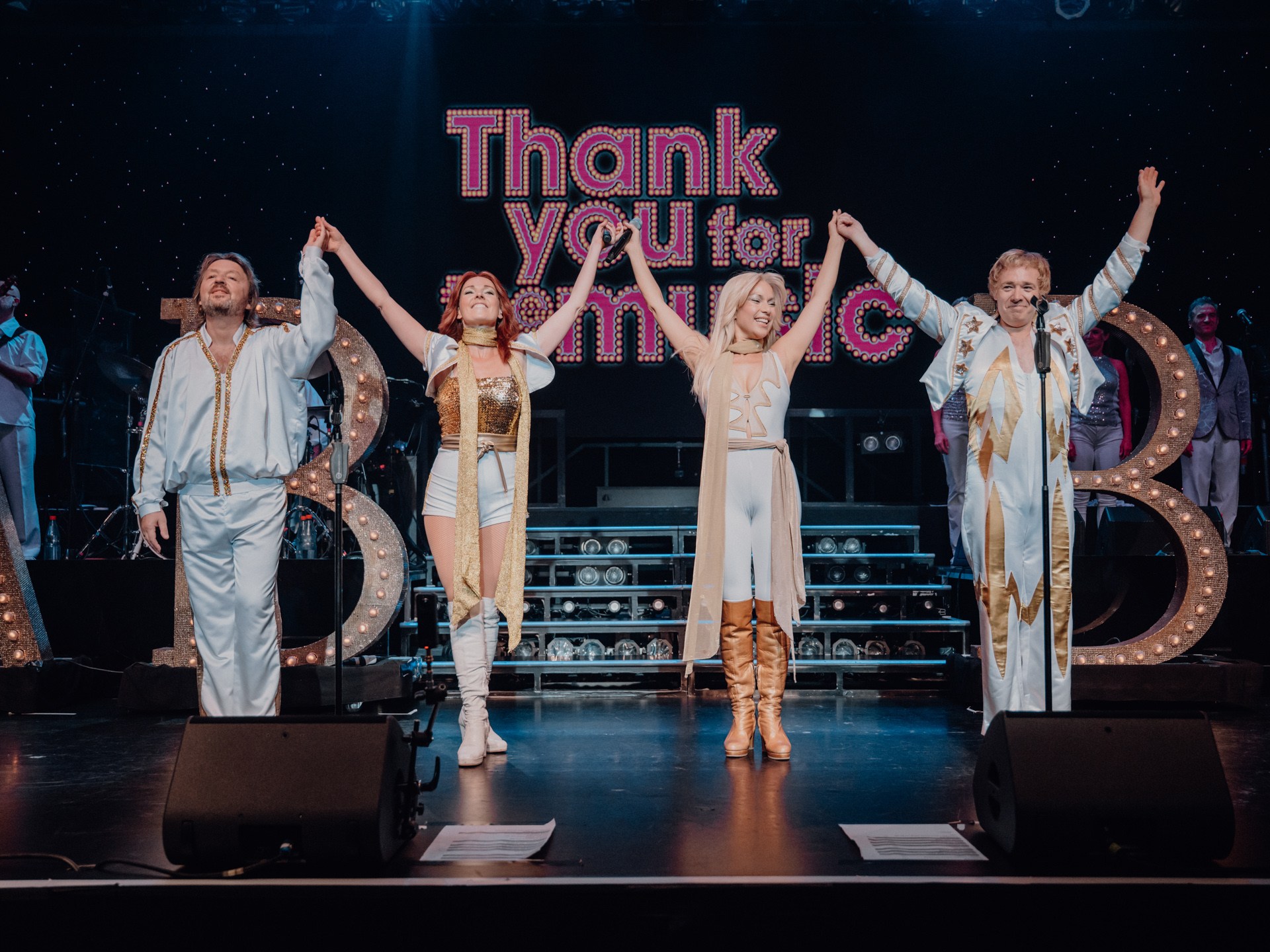 Four performers in white costumes hold hands on stage with 'Thank You for the Music' sign.