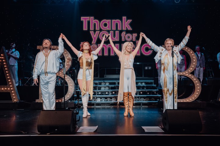 Four performers in white costumes hold hands on stage with 'Thank You for the Music' sign.