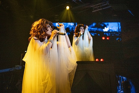 Performer in flowing gown sings passionately on stage with dramatic lighting.
