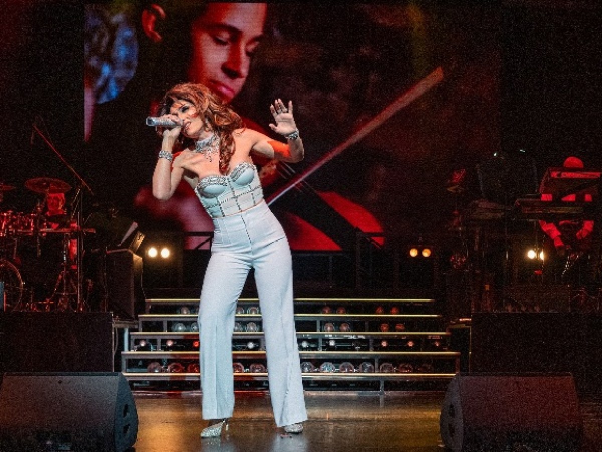 Singer performing energetically on stage with a band and large video screen in the background.