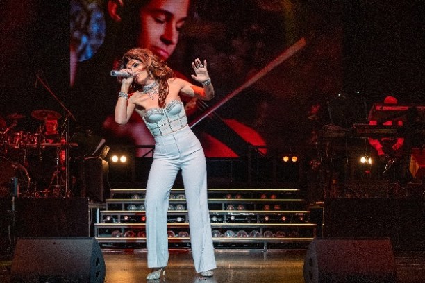 Singer performing energetically on stage with a band and large video screen in the background.